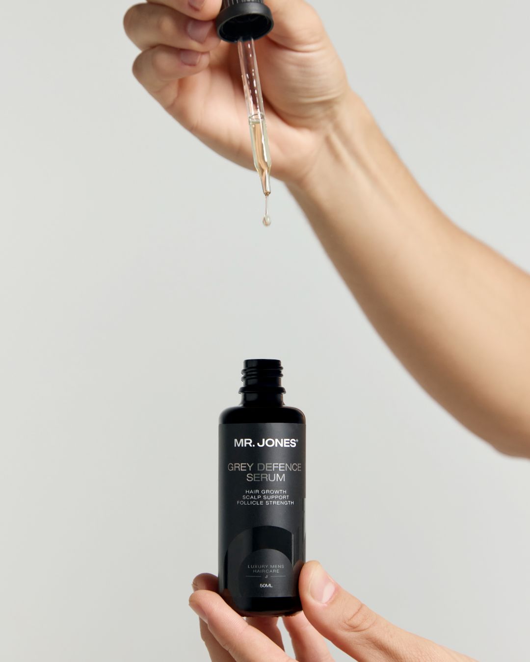 MR. JONES Grey Defence Serum bottle with dropper held by a hand against a light grey background