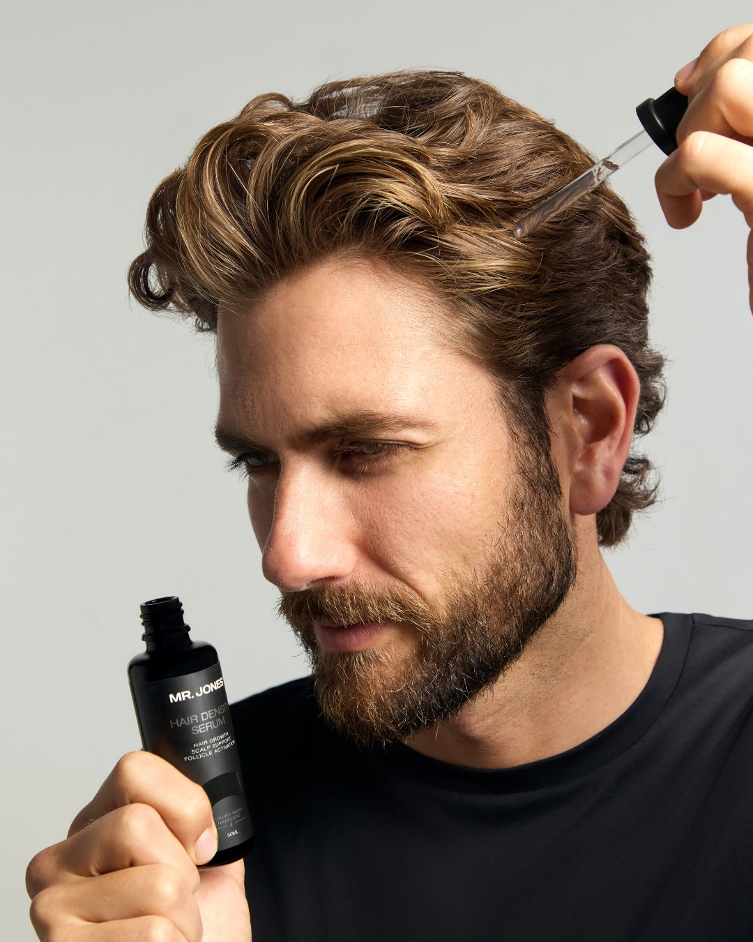 Man applying hair product 'Hair Density Serum' to his hair with a dropper bottle against a neutral background
