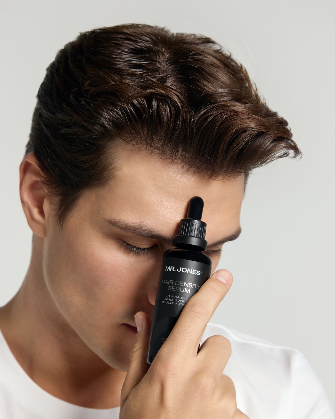 Man holding a black dropper bottle labeled 'Mr. Jones Hair Density Serum' close to his face against a plain background