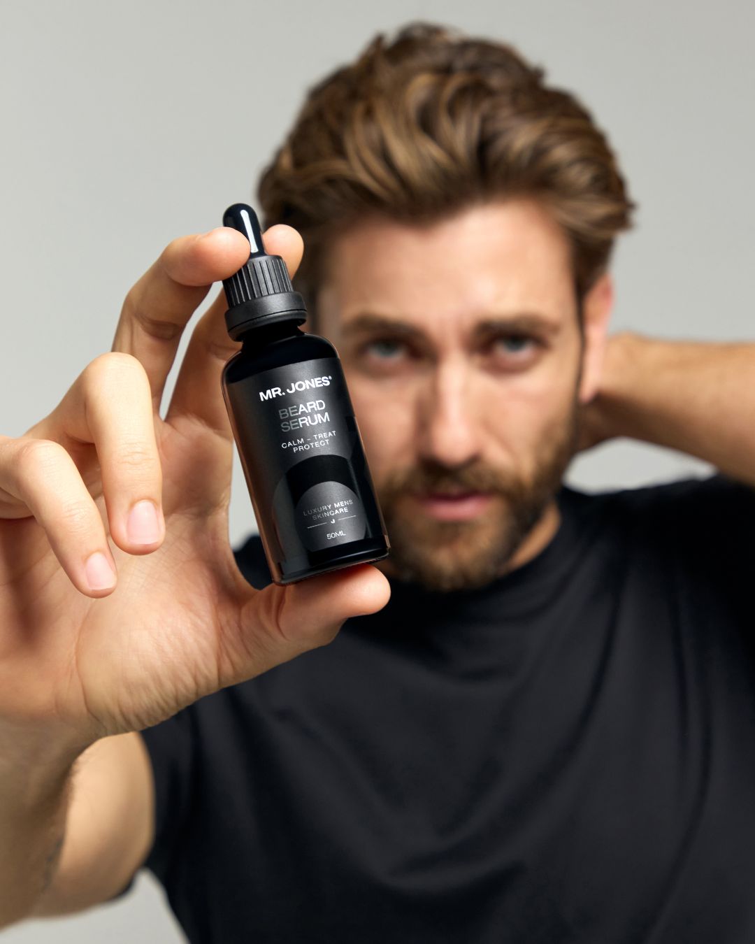 Man holding a black bottle with a dropper labeled 'Mr. Jones Beard Serum' against a neutral background